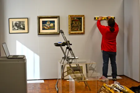 Three framed works on paper are hung on a gallery wall; to the right of them a person in a red shirt faces the wall holding a level. A steplader is visible in the foreground.