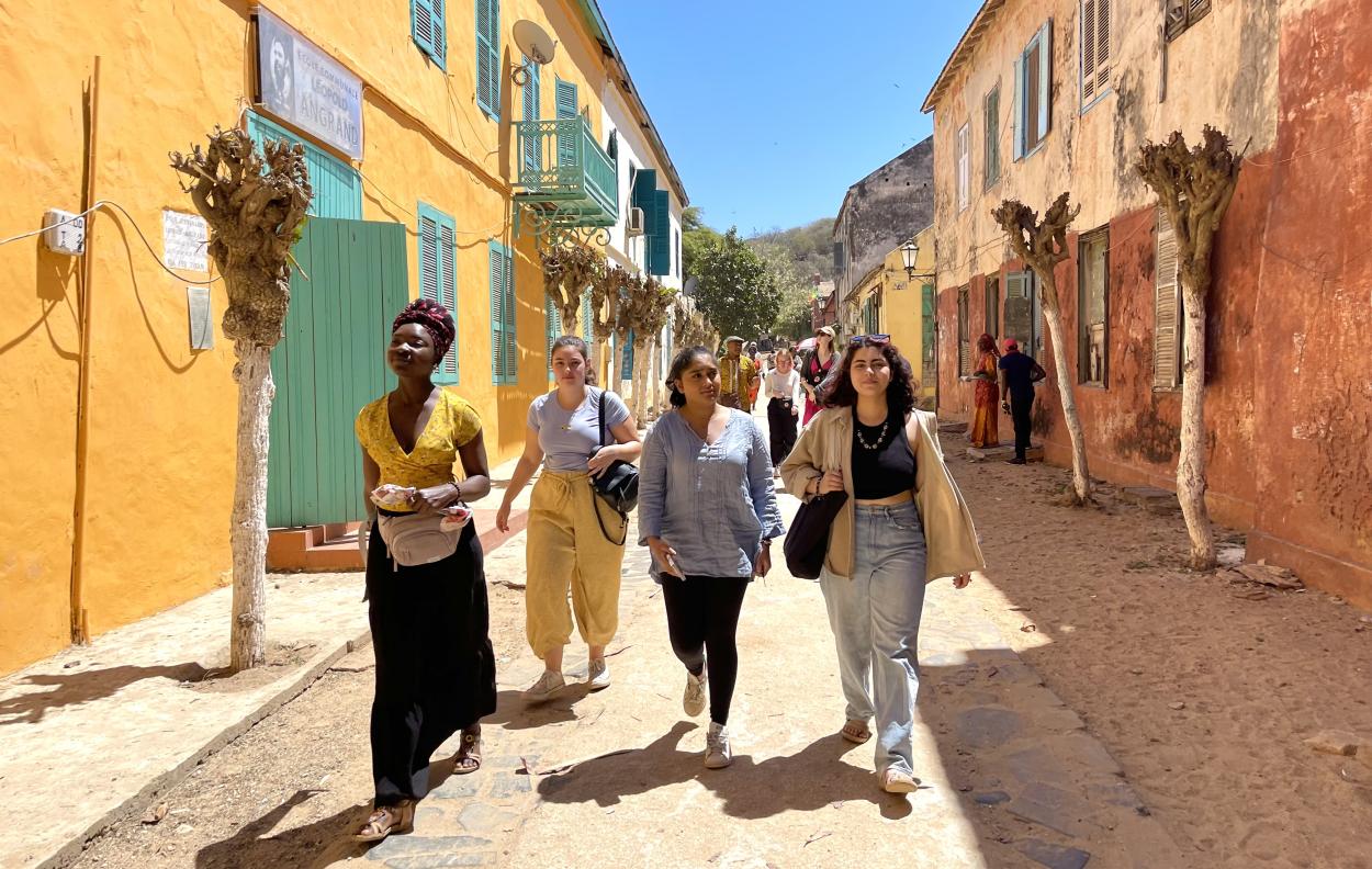 Four students walk down a dirt road with brightly colored two-storey buildings on either side.
