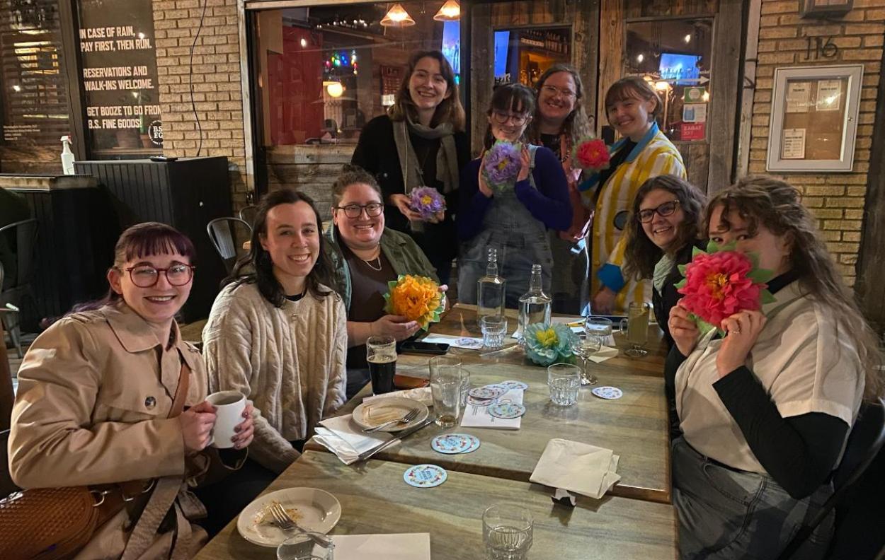 A group of Mawrters smile together at a restaurant table. They hold paper flowers.