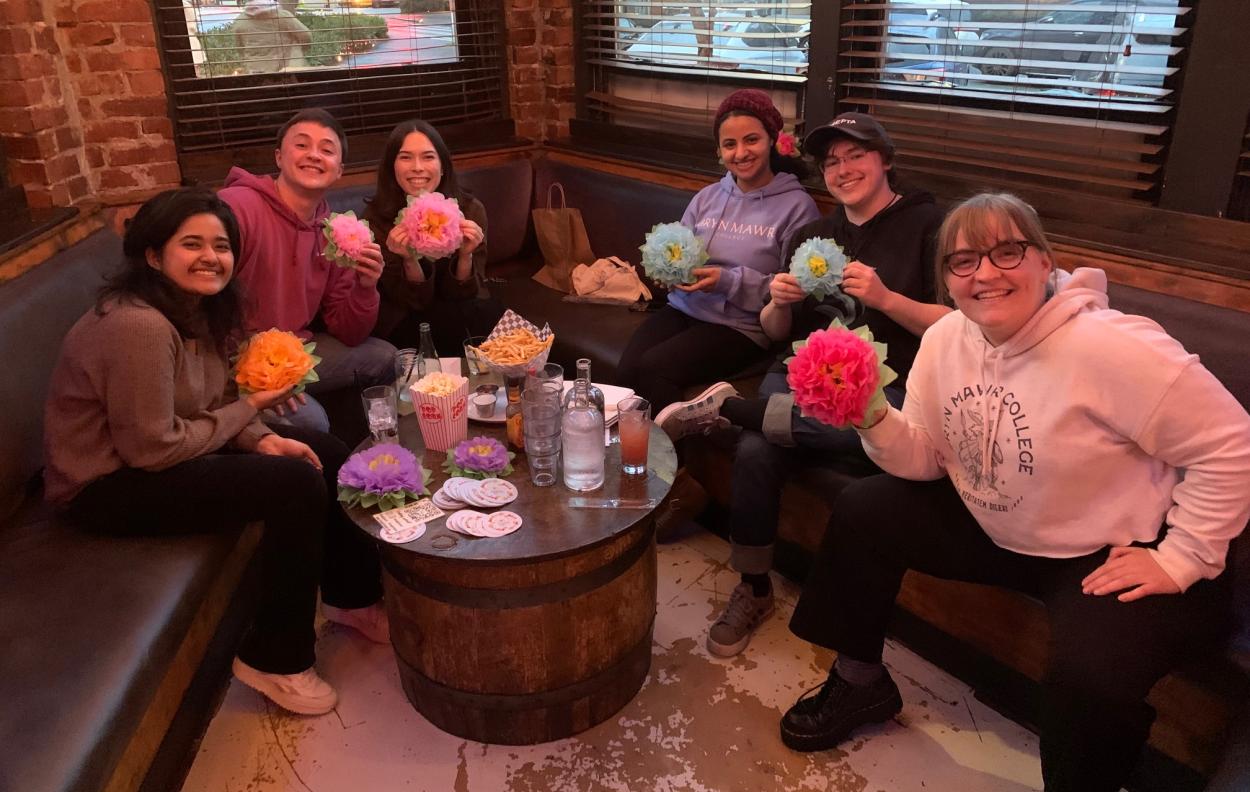 A group of Mawrters smile together at a restaurant table. They hold paper flowers.