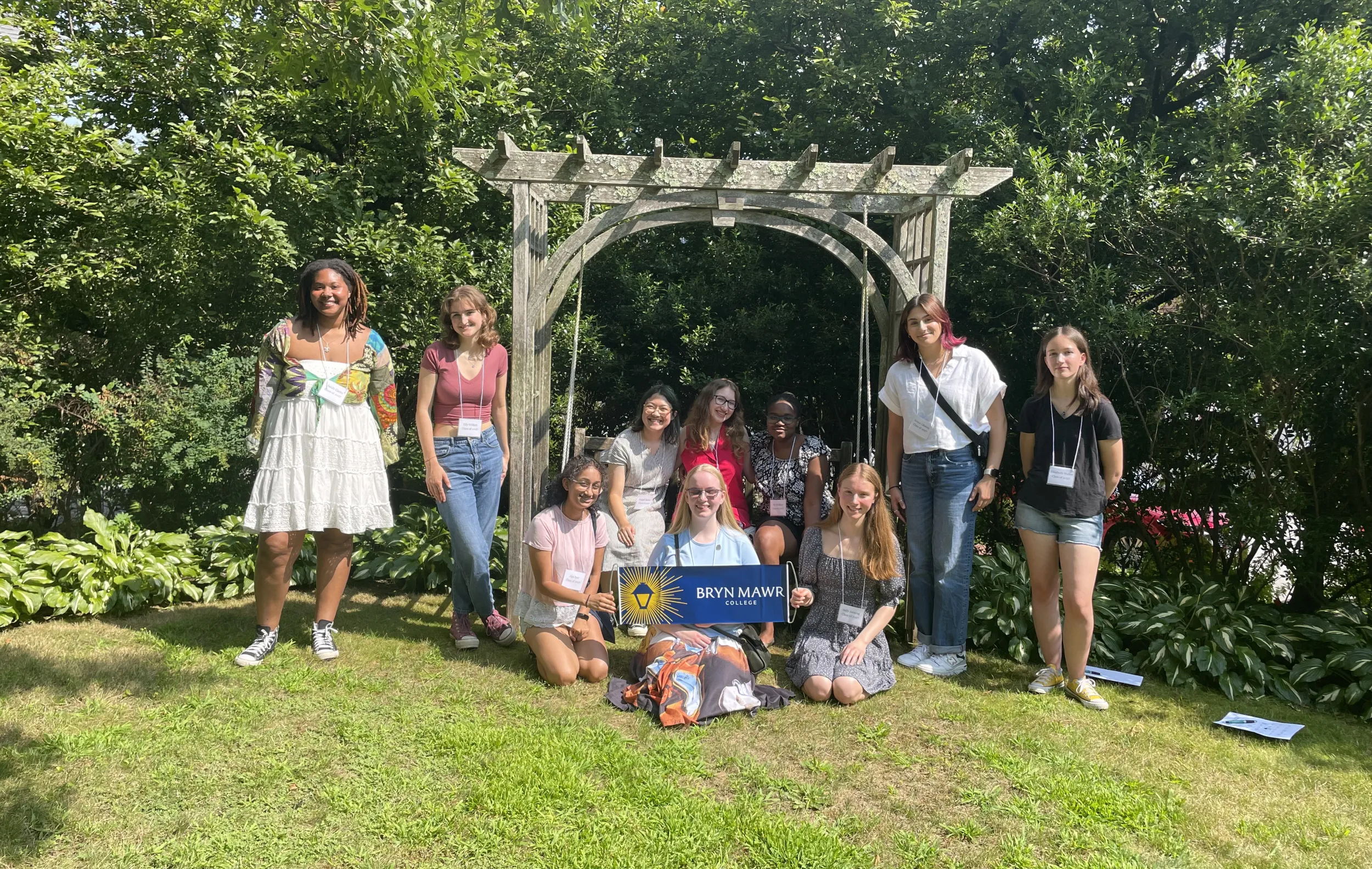 10 students, some sitting and some standing, around a wooden swing on a green lawn