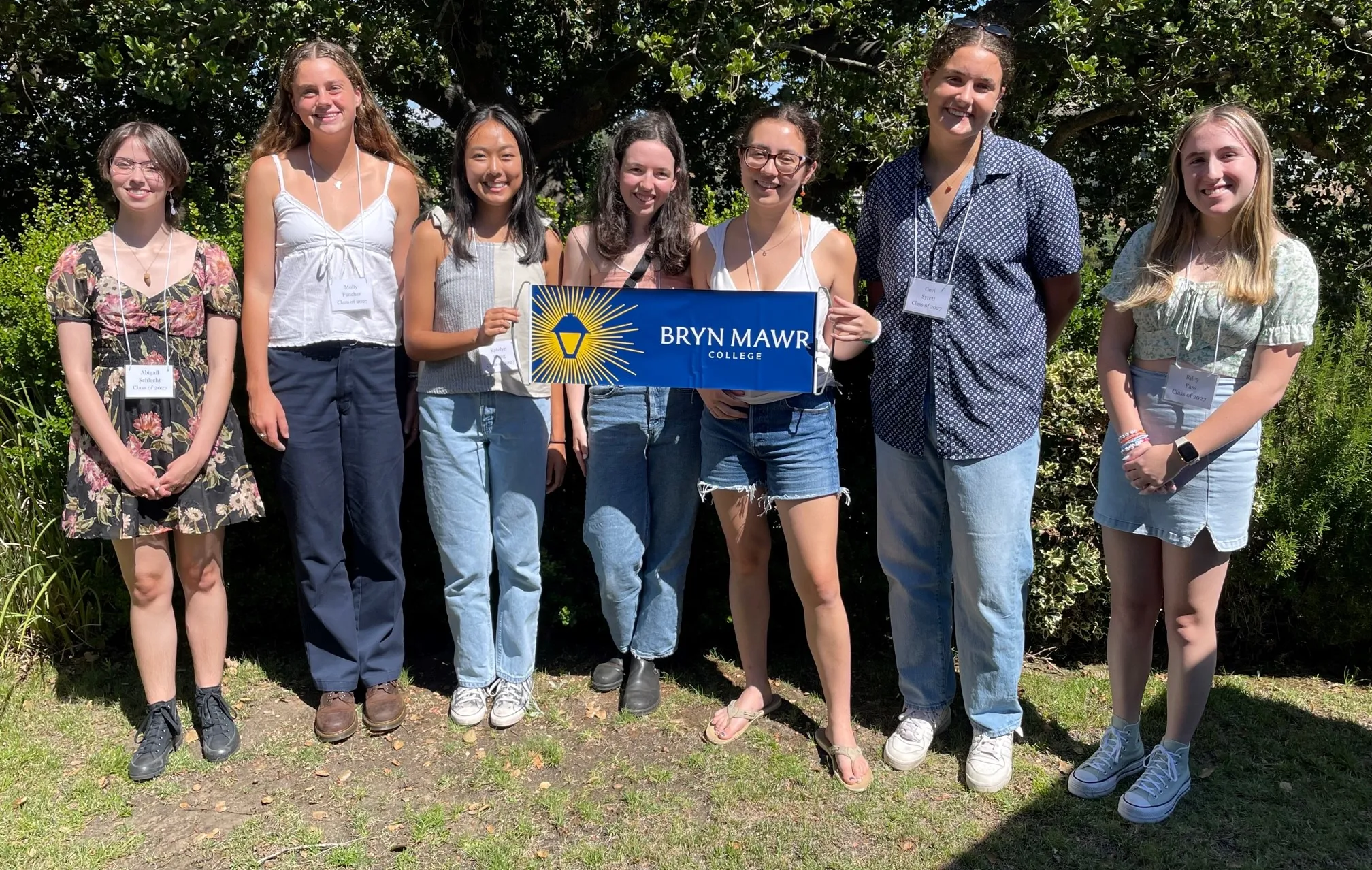 Seven students holding a Bryn Mawr College banner stand in the sunshine, smiling at the camera