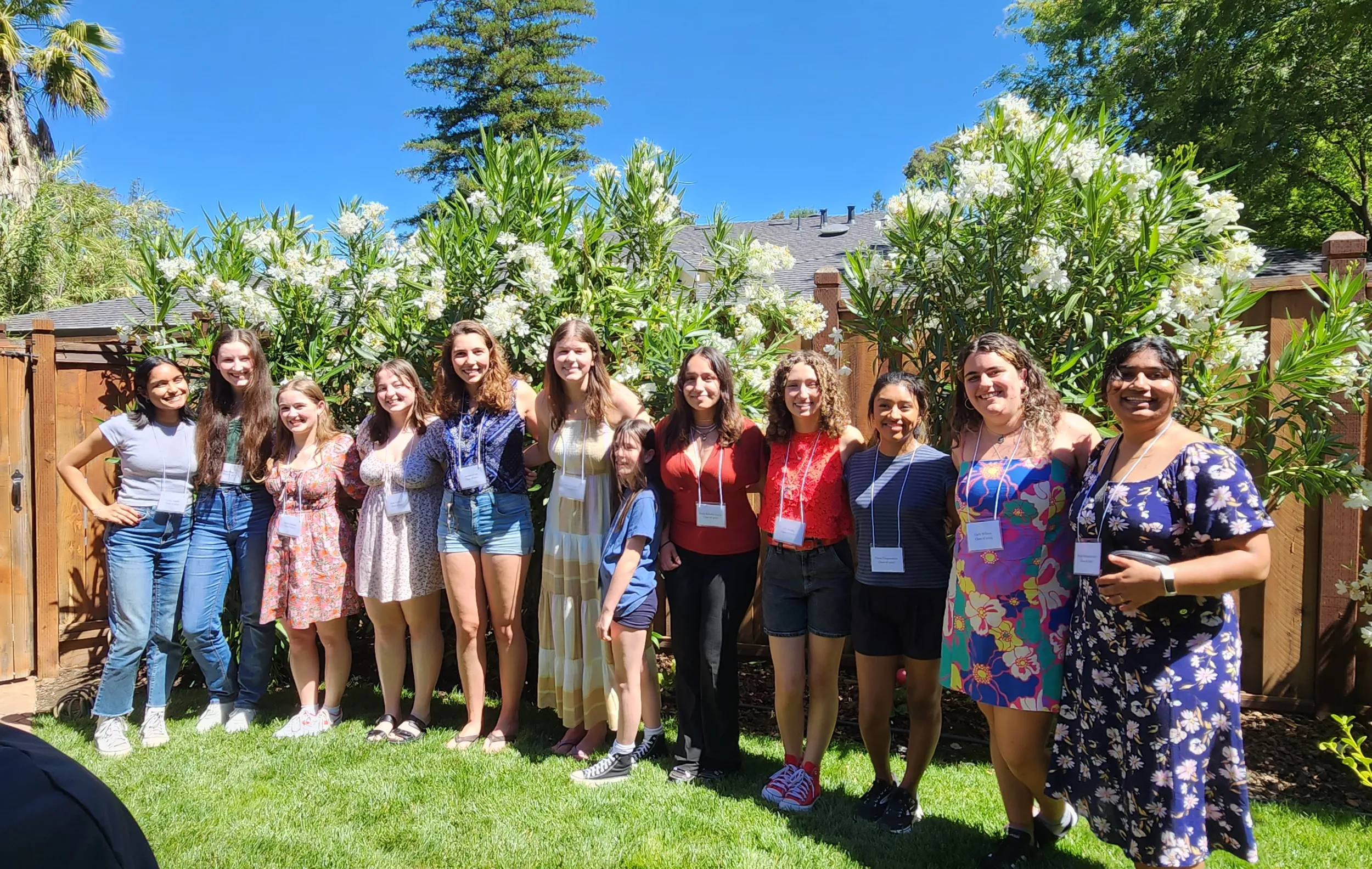 A group of students stand in the sunshine in front of a wooden fence. They're smiling at the camera.