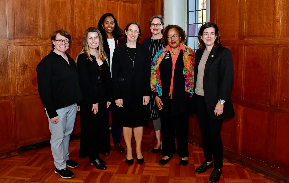 Seven women of different ages stand in front of a wood paneled wall and smile at the camera