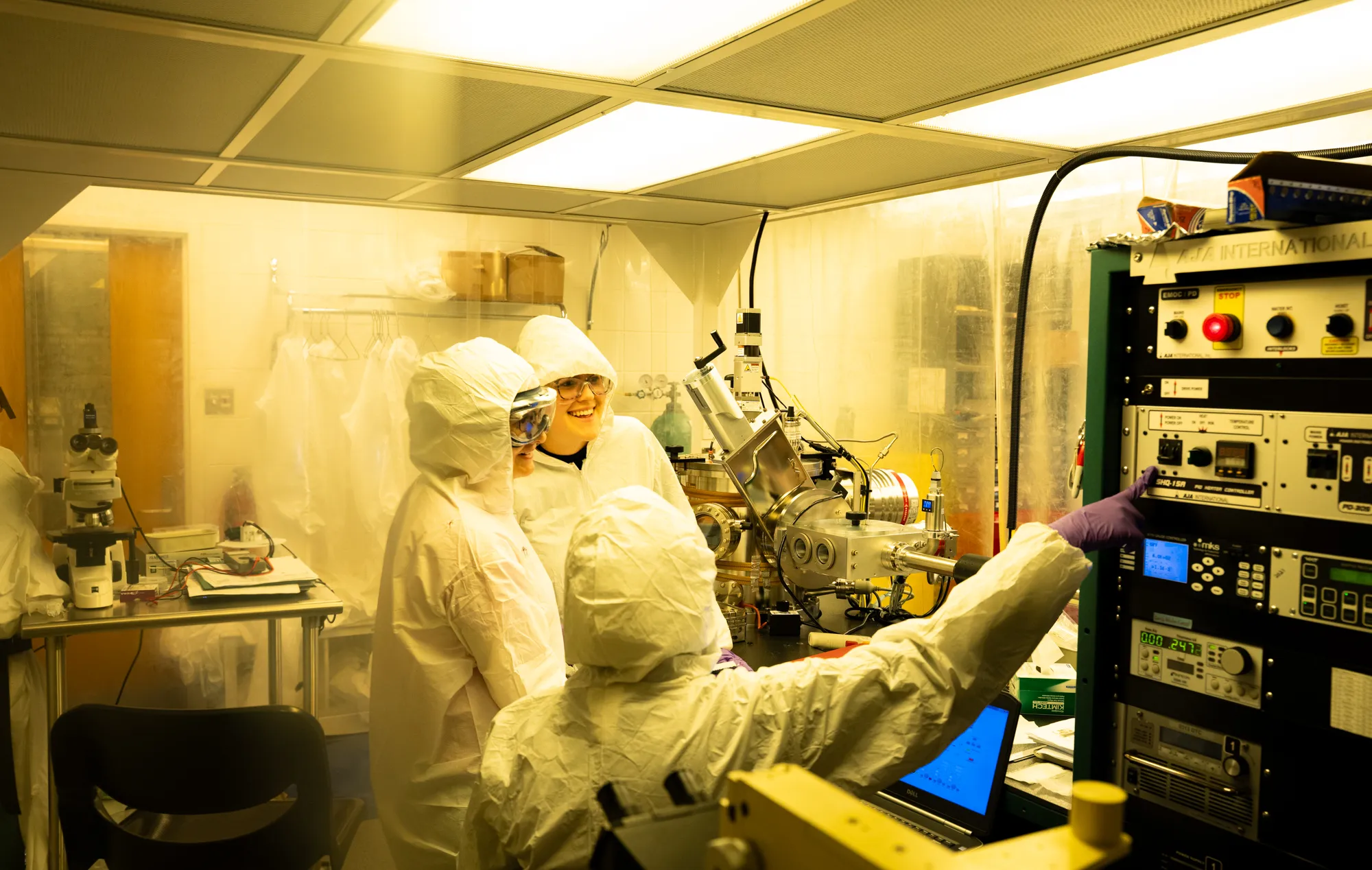 Three researchers in head to toe protective gear work in a laboratory with large black rectangular machine on the right.