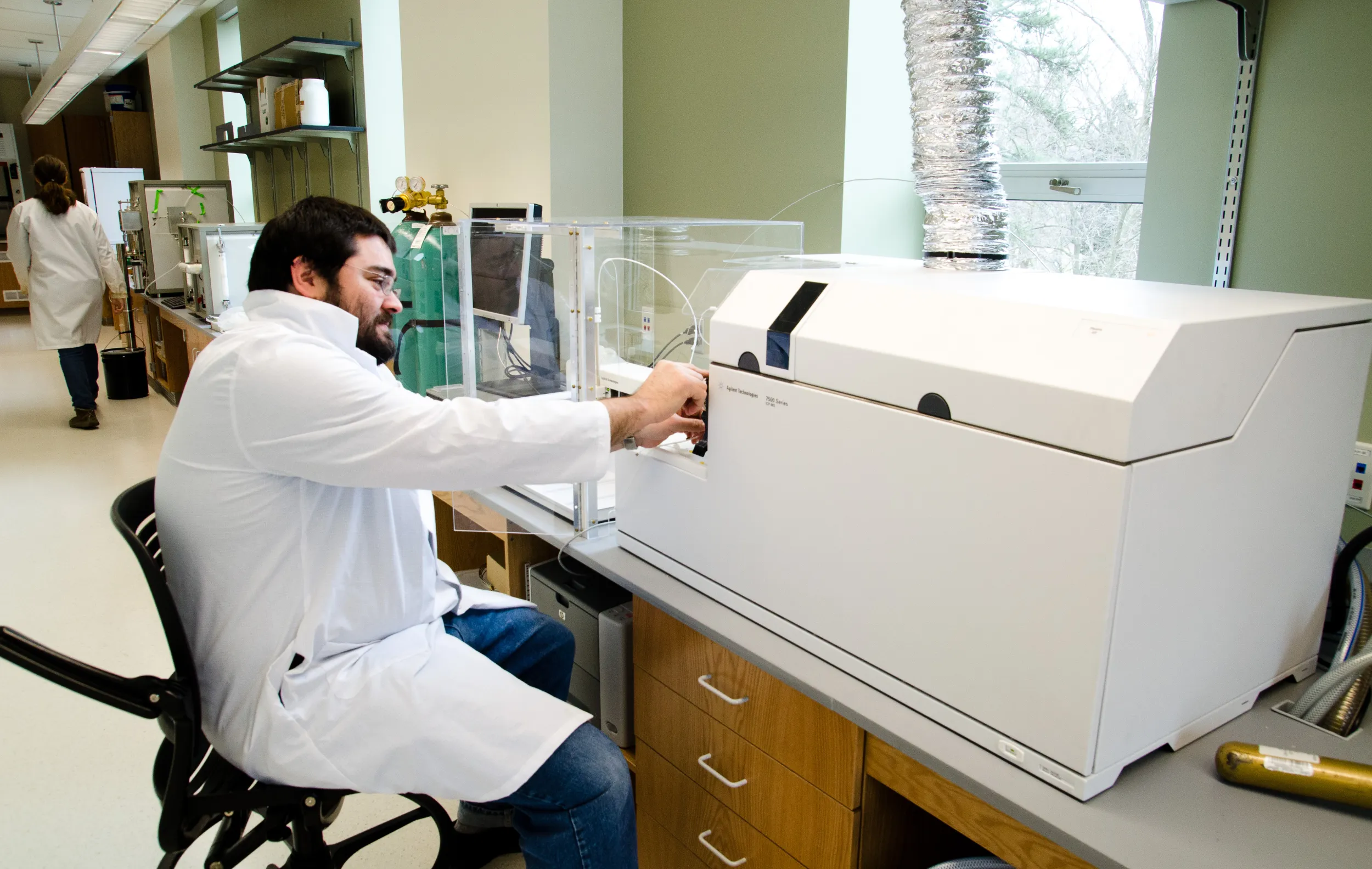 A man with light skin, dark hair and a beard sits at a piece of mineral analysis equipment