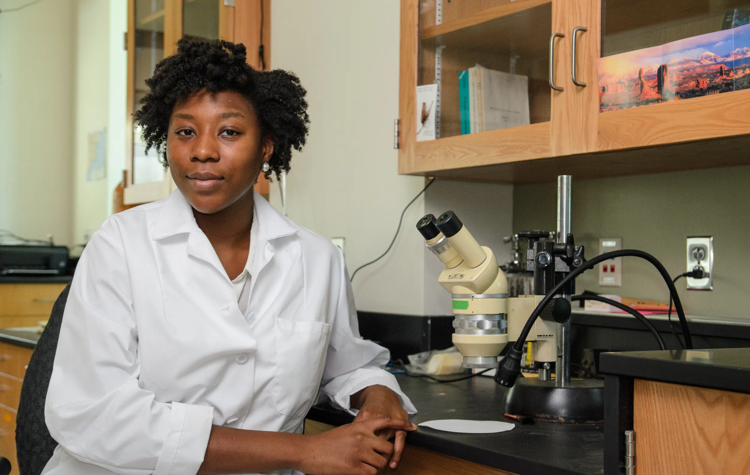 A young woman with dark skin and hair wearing a white lab coat sits posted for a portrait next to microscopes and other equipment in a science lab 
