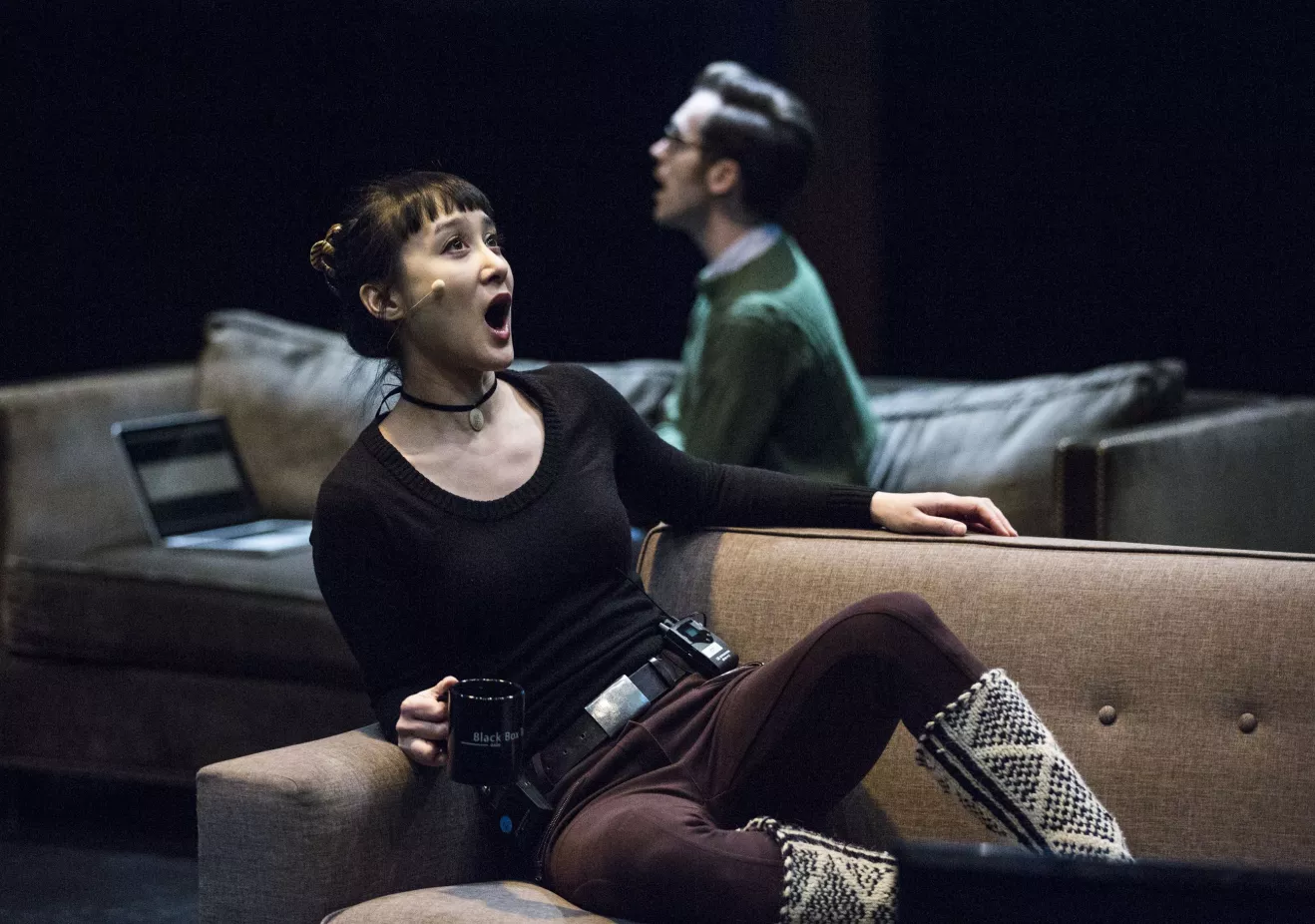A woman in the foreground sits on a couch facing right and sings. A man in the background on a couch faces left.