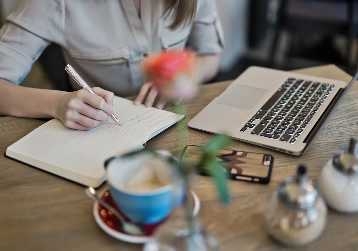 Person writing with a laptop, phone, coffee, and plant