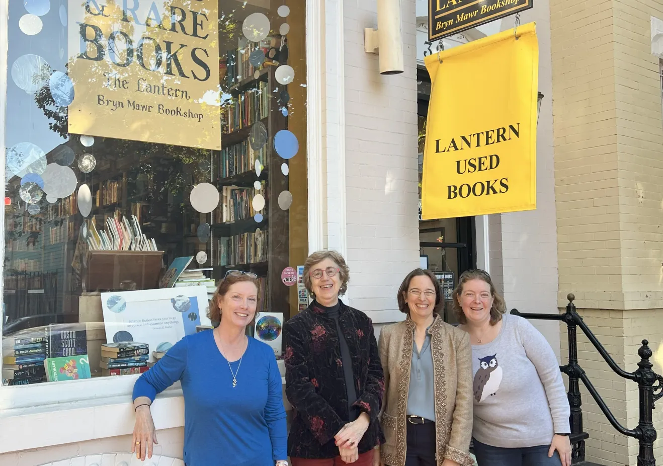 Four women stand and smile in front of a white brick building.