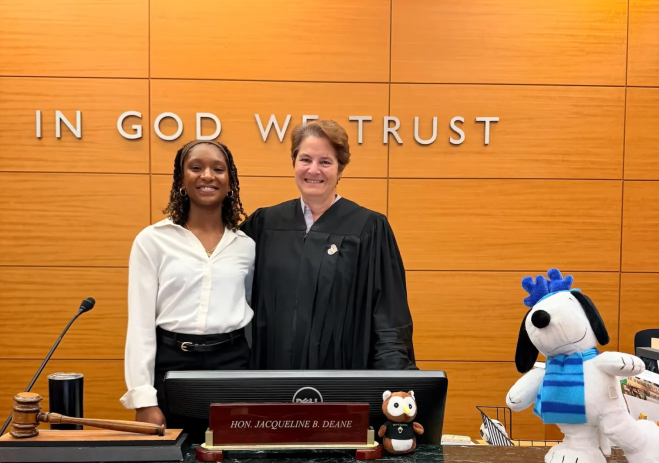 Two women, one of whom is wearing judicial robes, smile at a camera in front of a wall that says "In God We Trust"