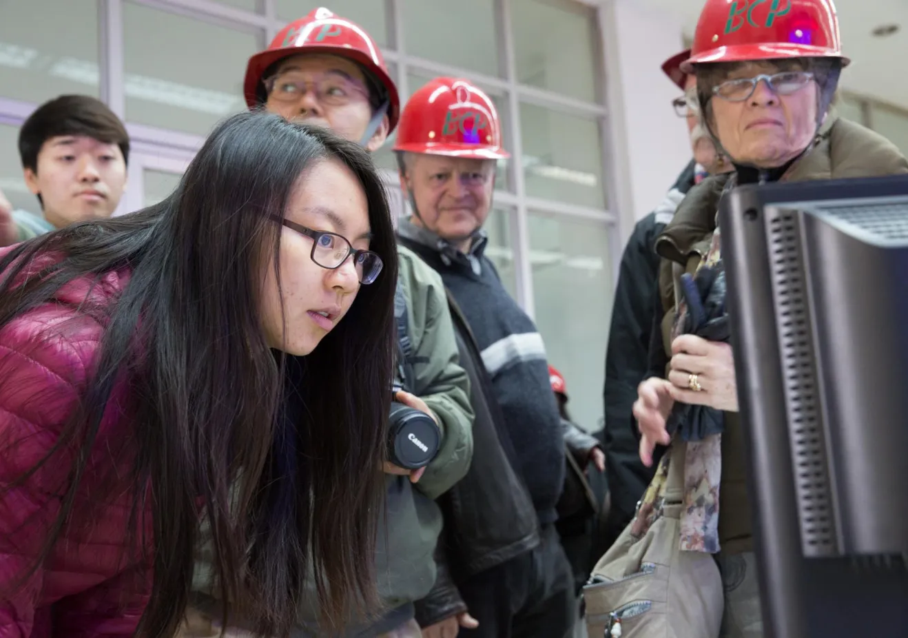 A young woman with long black hair and glasses looks at a computer screen while several people in red hard hats stand behind her 