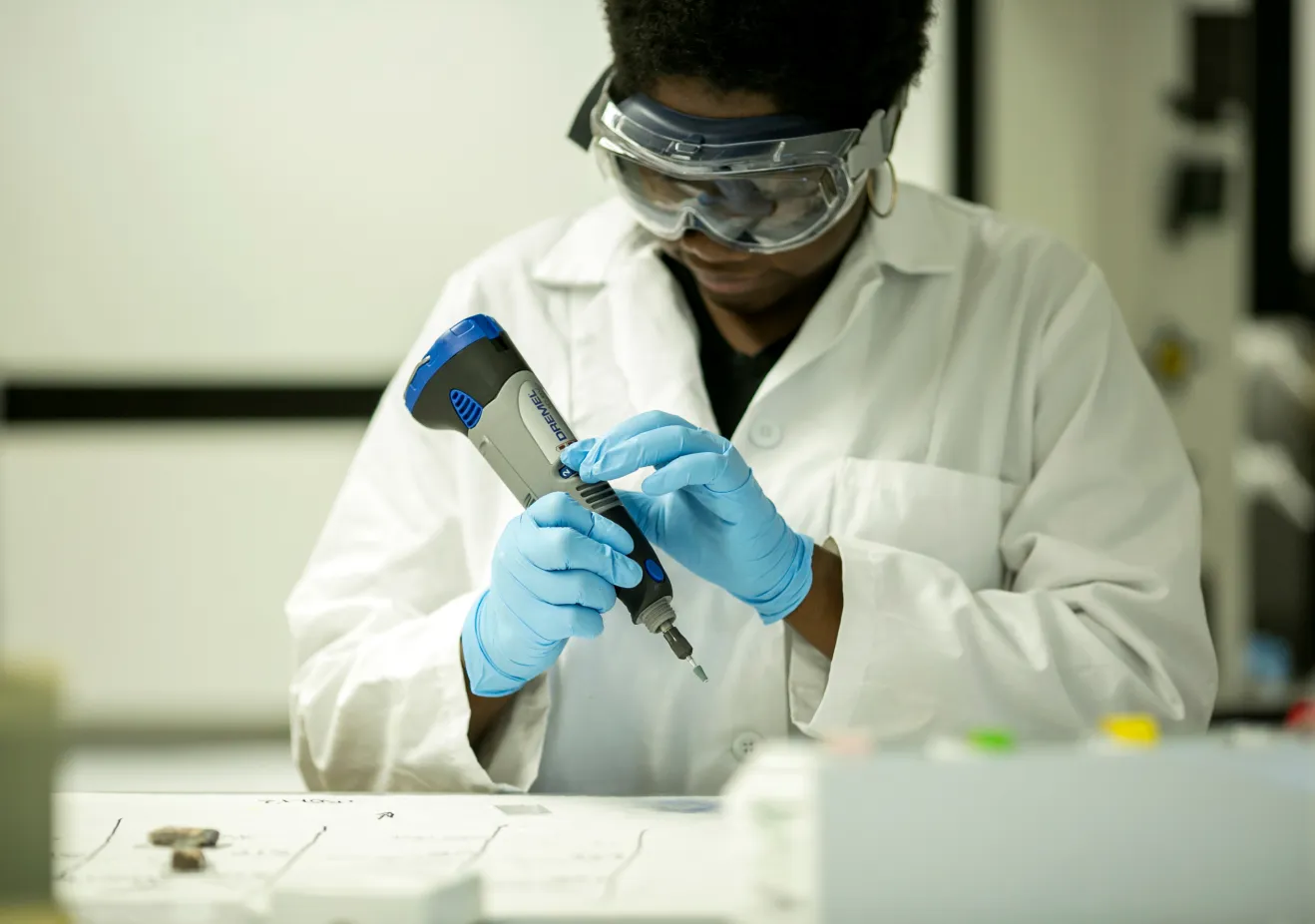 a student with dark skin and short curly black hair wearing protective gear including a lab coat, googles, and gloves works with geology samples in a lab