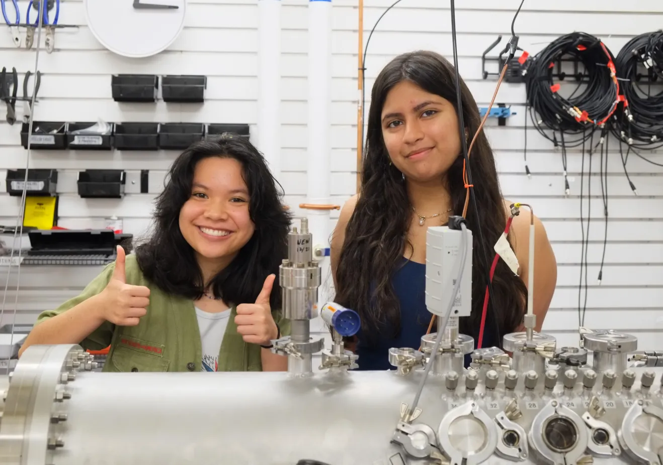 Two students with tan skin and long dark hair stand behind a piece of plasma science equipment smiling, while the one of the left gives two thumbs up