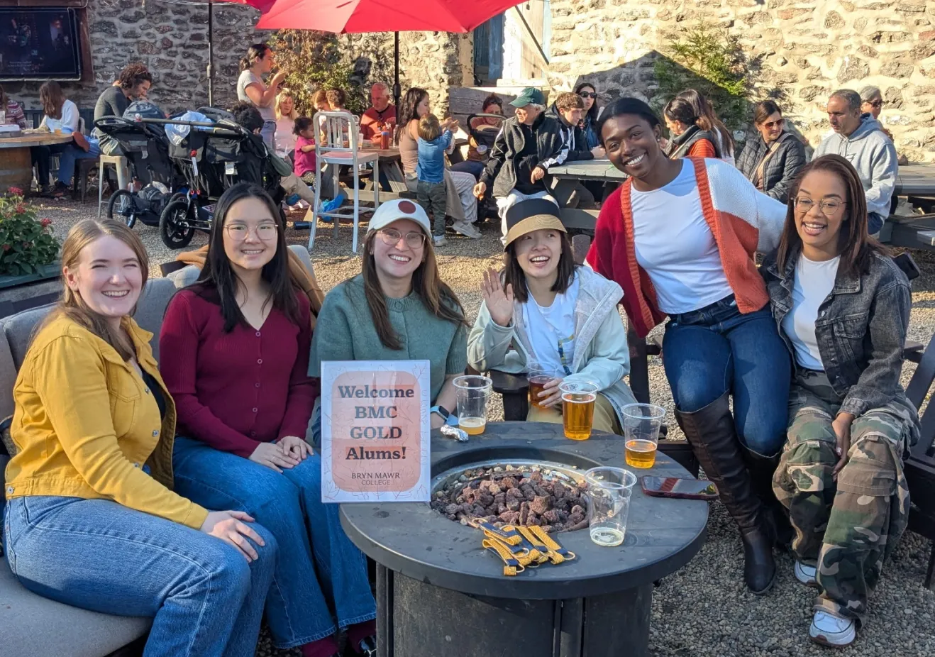 A group of young women sit around a fire pit and smile at the camera