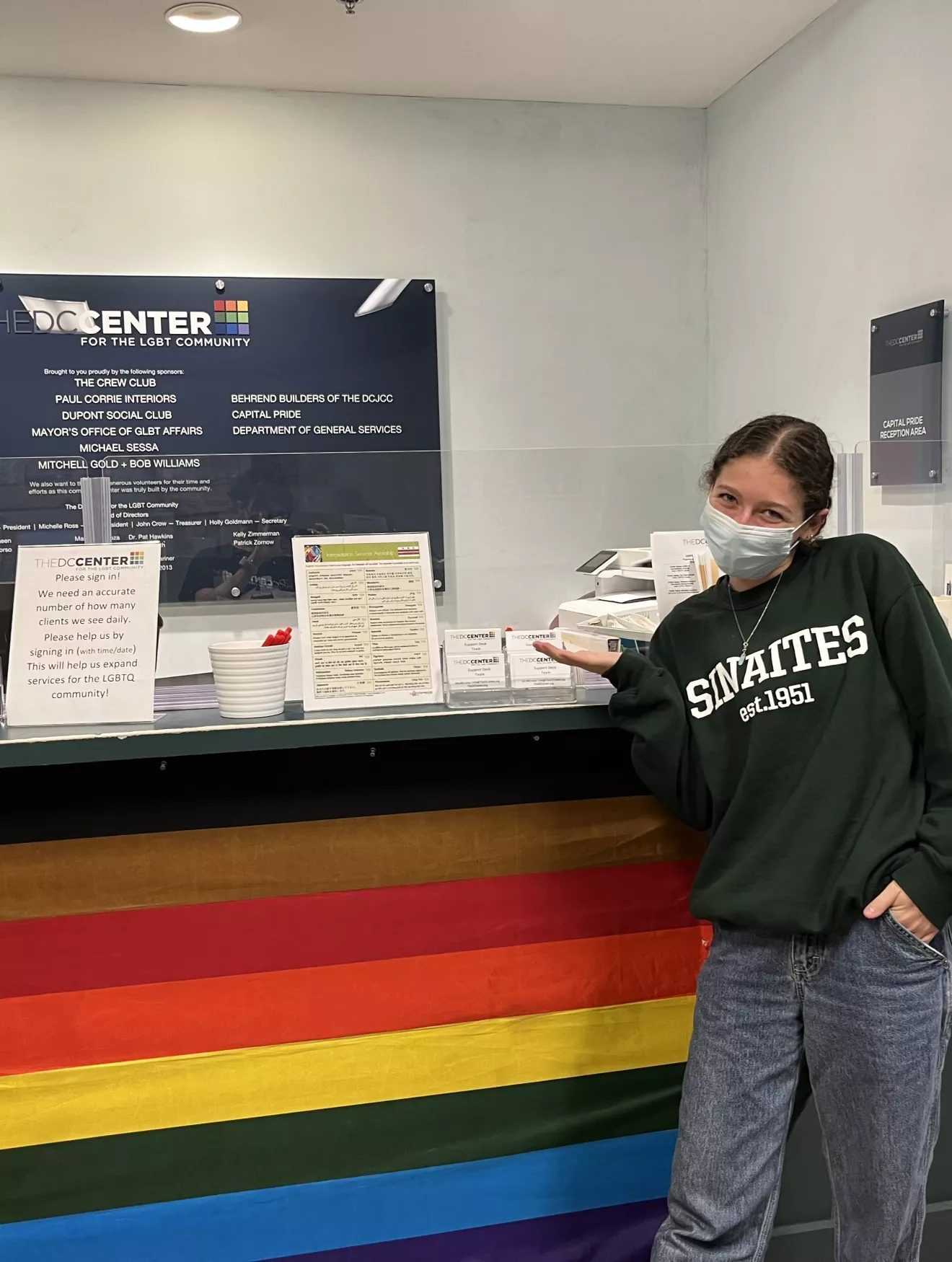Elana standing in front of a desk with a Rainbow flag
