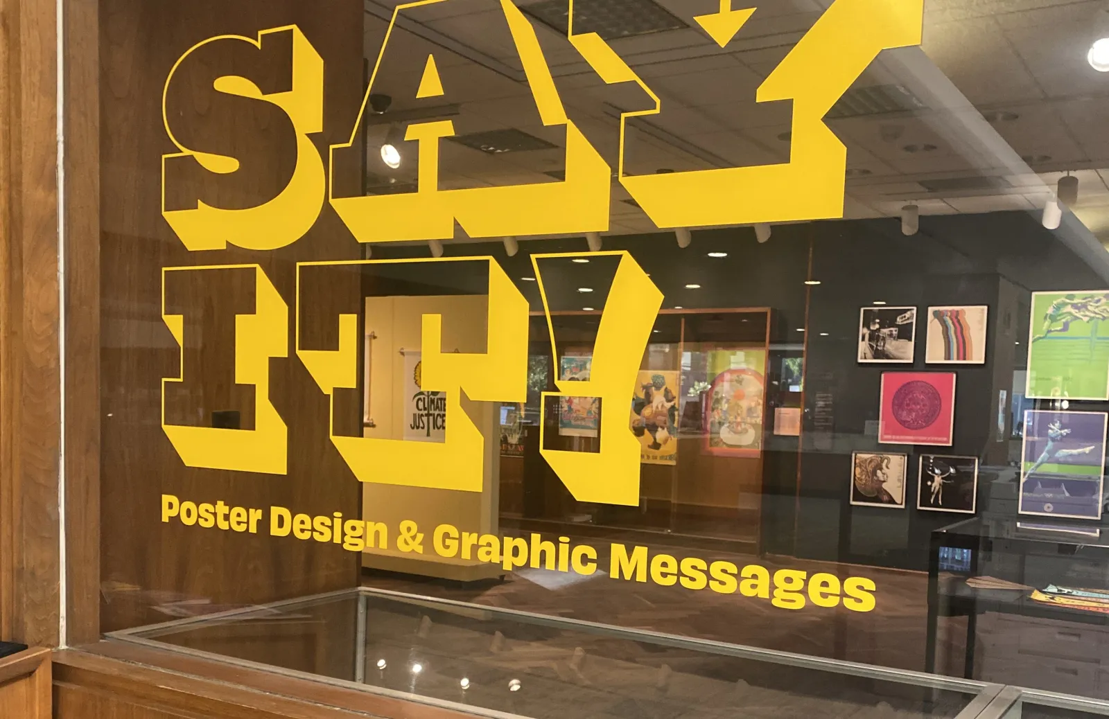 SAY IT! Poster Design & Graphic Messages is written in yellow vinyl on the window of the 1912 Gallery. Through the window you can see multicolored posters hanging on the wall.