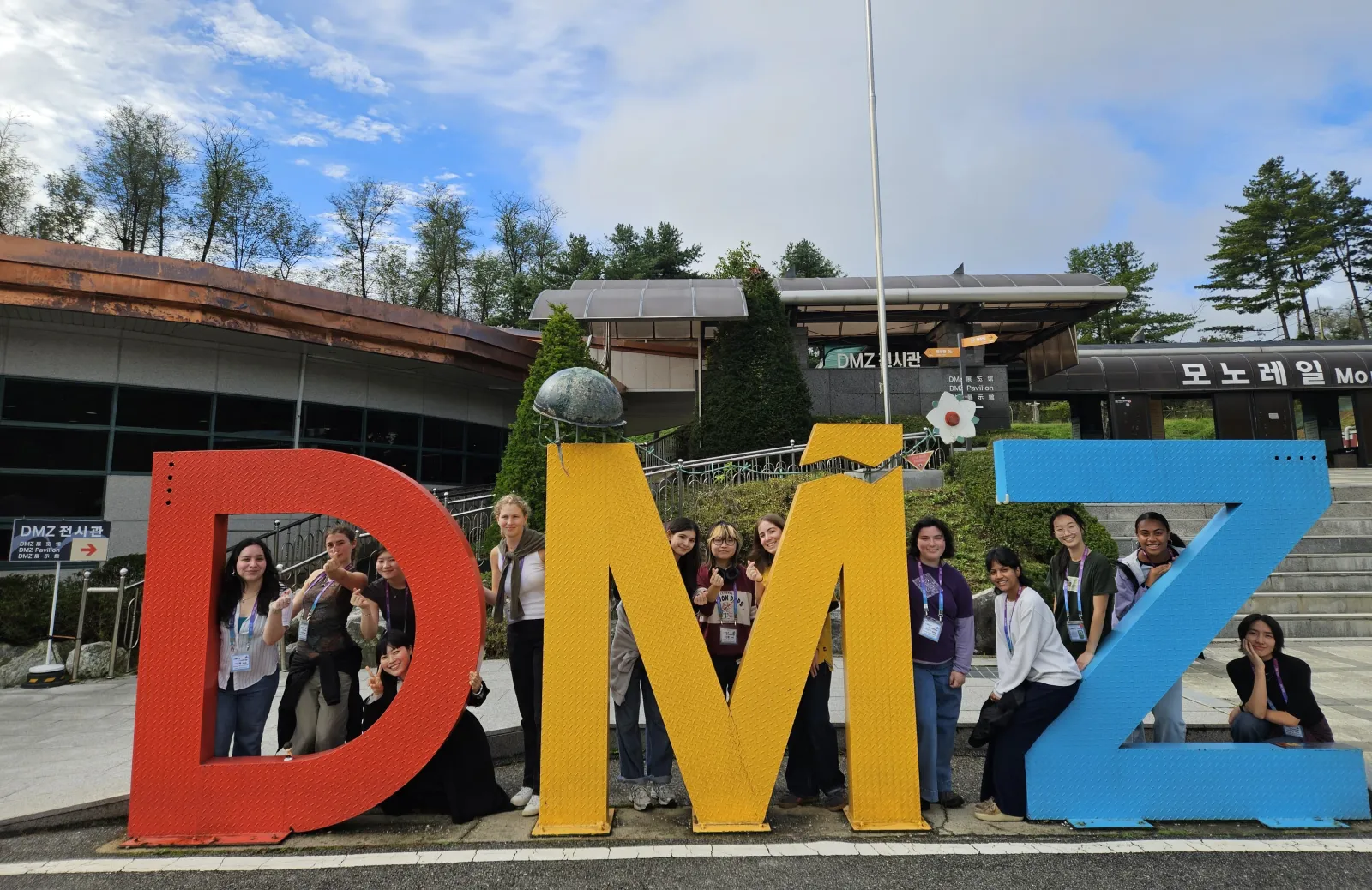 12 students behind a sculpture depicting the letters "D" "M" "Z" in primary colors.