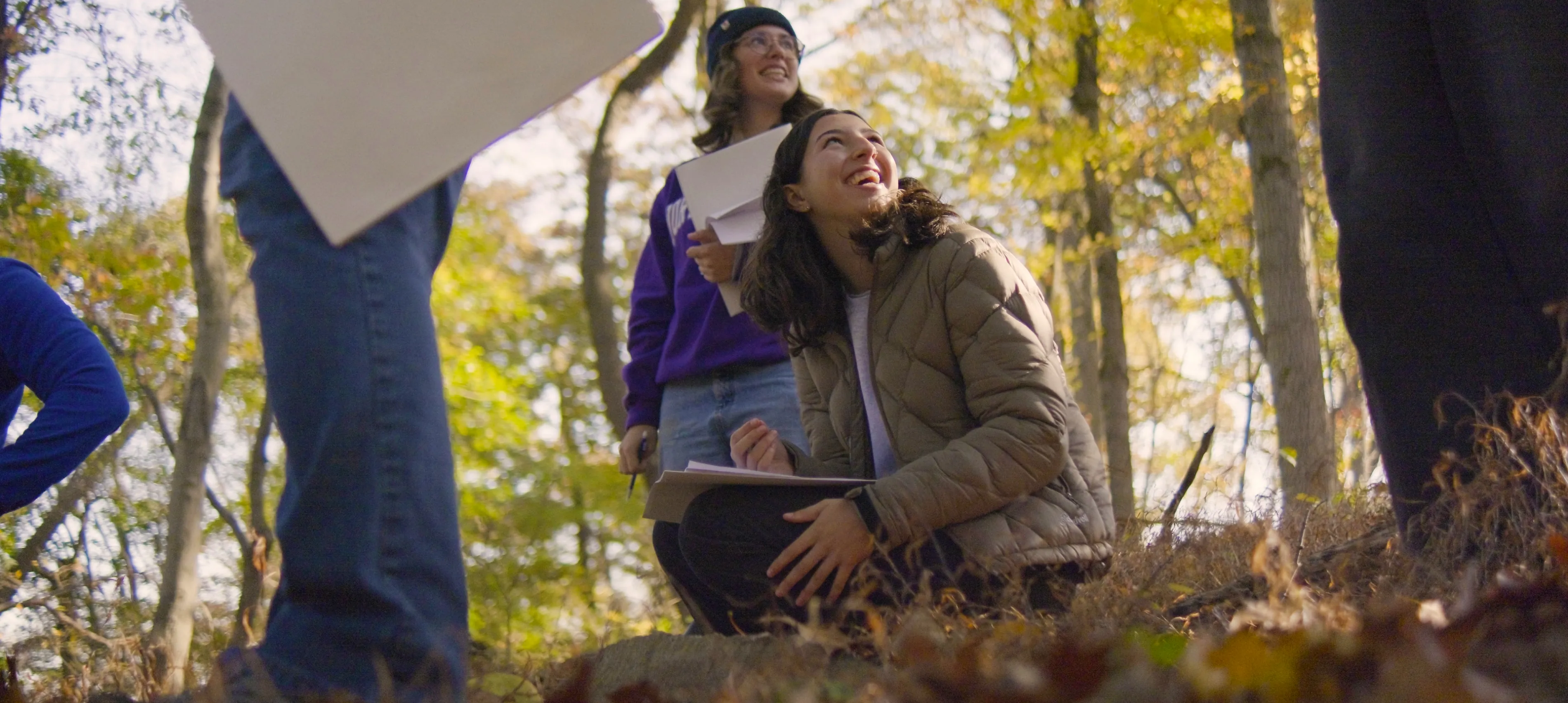 A group of students stands in a forest in autumn with one student crouching in the center and looking upwards