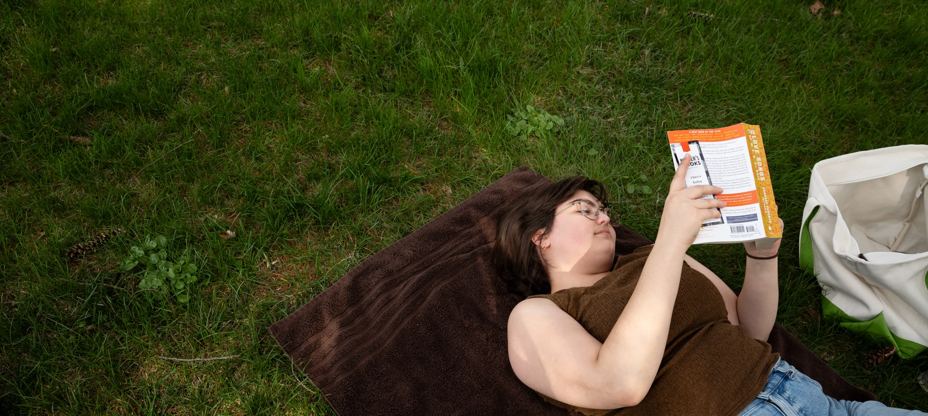 A student reads a book while lying on a towel on the grass