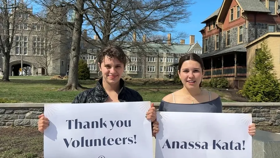 Two students stand outside holding signs that say "Thank you, volunteers!" and "Anassa Kata!"