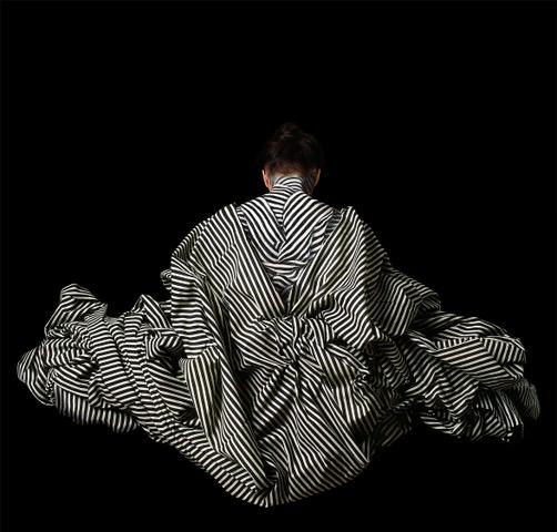 Cecilia Paredes in Black and White Striped garment against black ground, facing away.