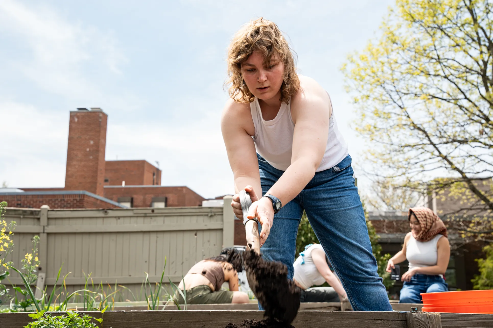 A young woman in a white tank top and blue jeans digs in a garden