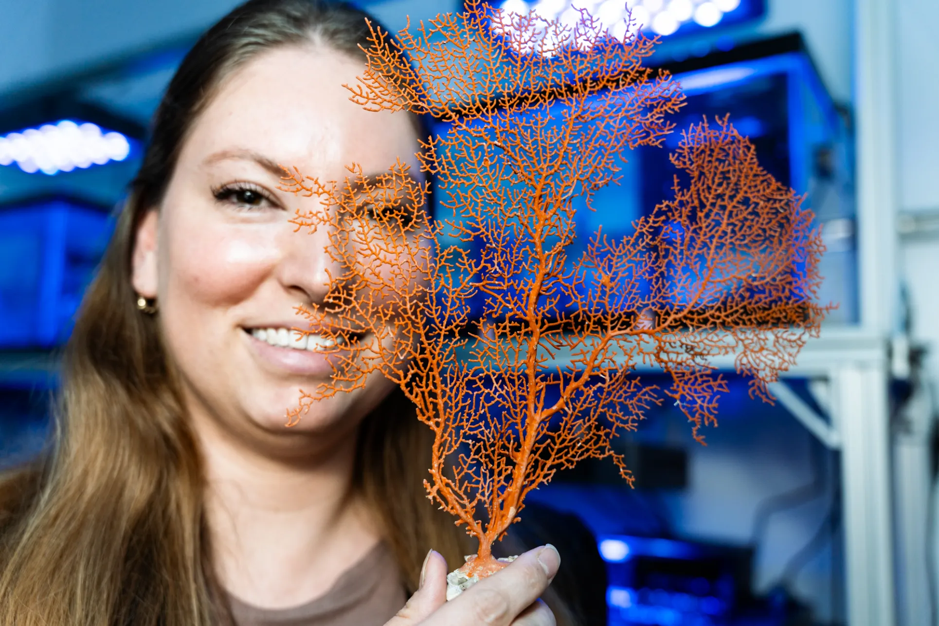 A woman with light skin and long brown hair holds a piece of orange coral in front of her face in a laboratory lit in blue