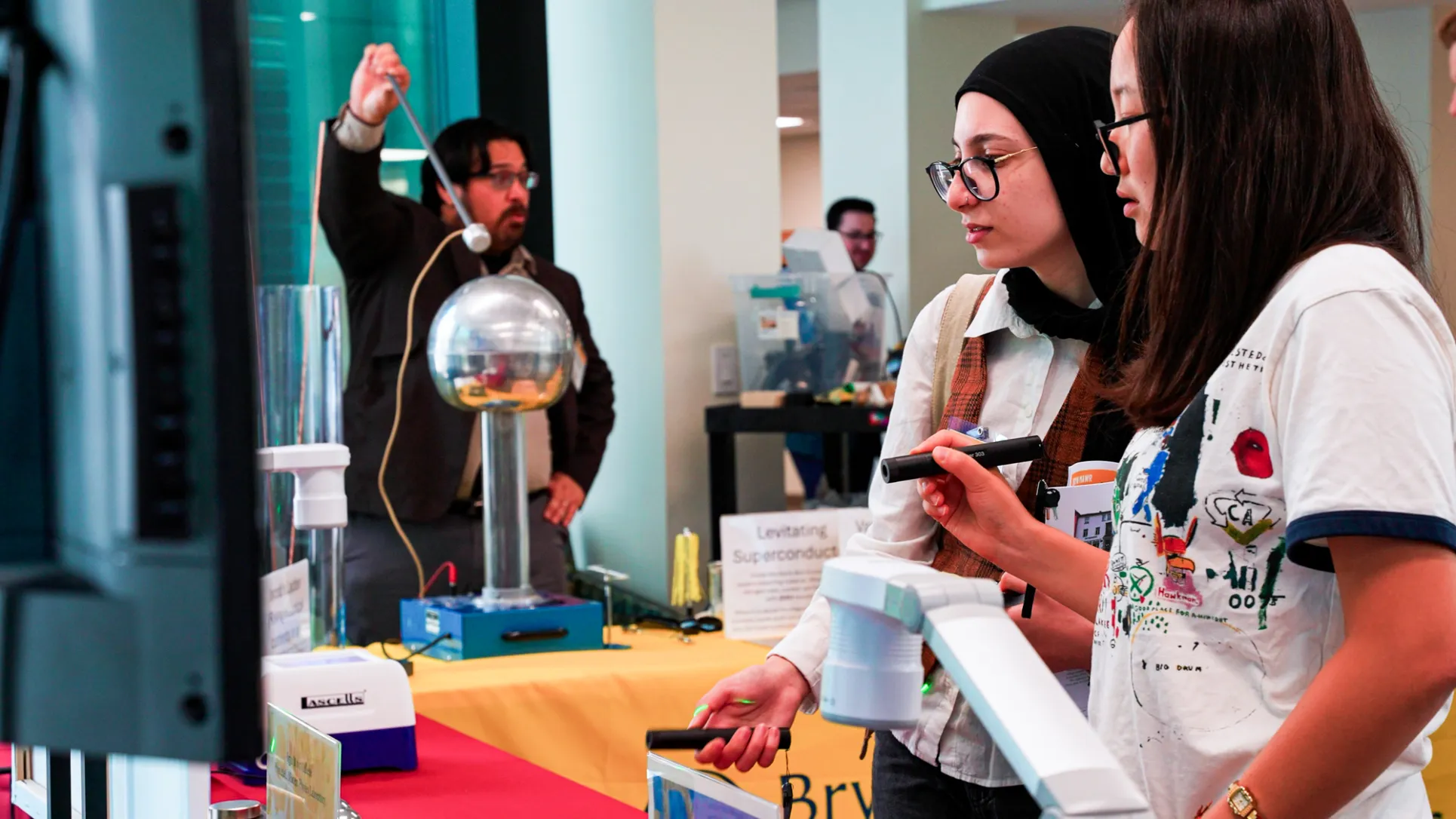 Two students with glasses, one with long dark hair and the other wearing a hair covering use laser pointers while a professor with a beard demonstrates something with a large sphere in the background
