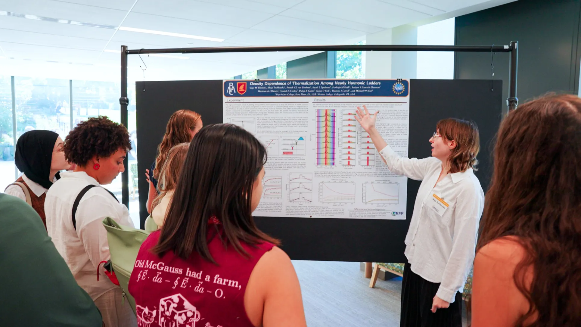 A student with light skin and medium length wavy red hair presents physics findings to a group of onlookers