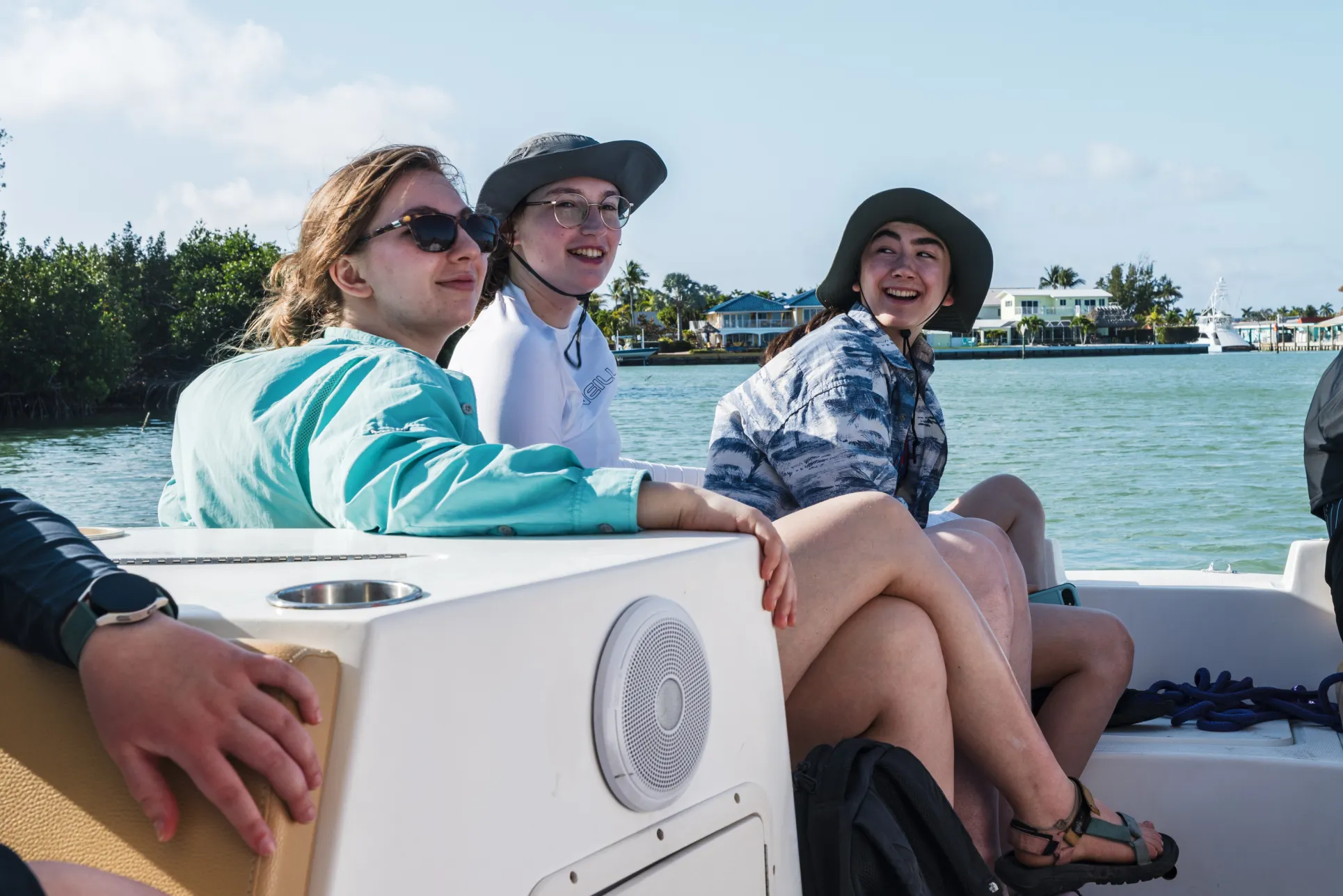 Three young people with long hair and light skin wearing swimming and snorkeling apparel sit on a boat in a clear lagoon