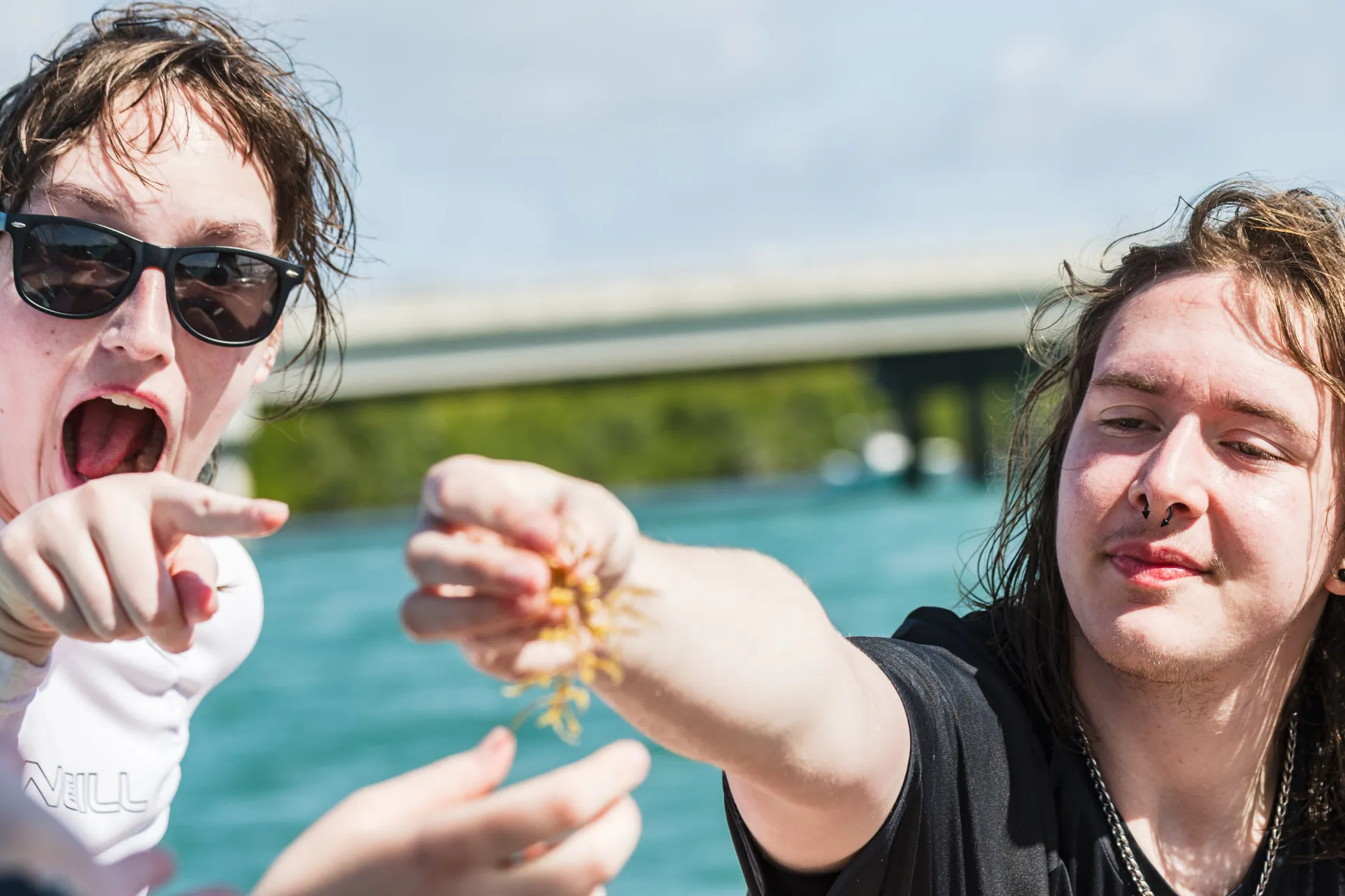 Two students with long hair and light skin stand in a blue lagoon. The student on the right holds up a specimen found in the water and the student on the left points with an expression of surprise