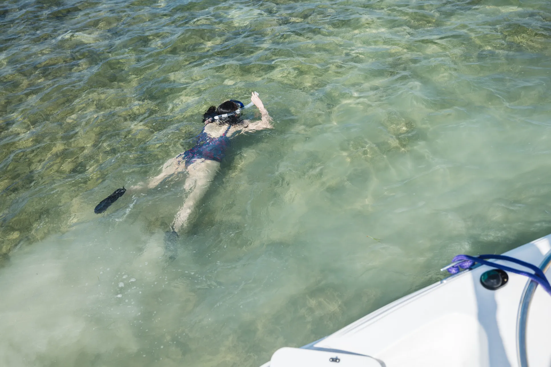 Woman in a one piece blue toned swimsuit snorkels in clear water, with the hull of a boat seen next to her as from above