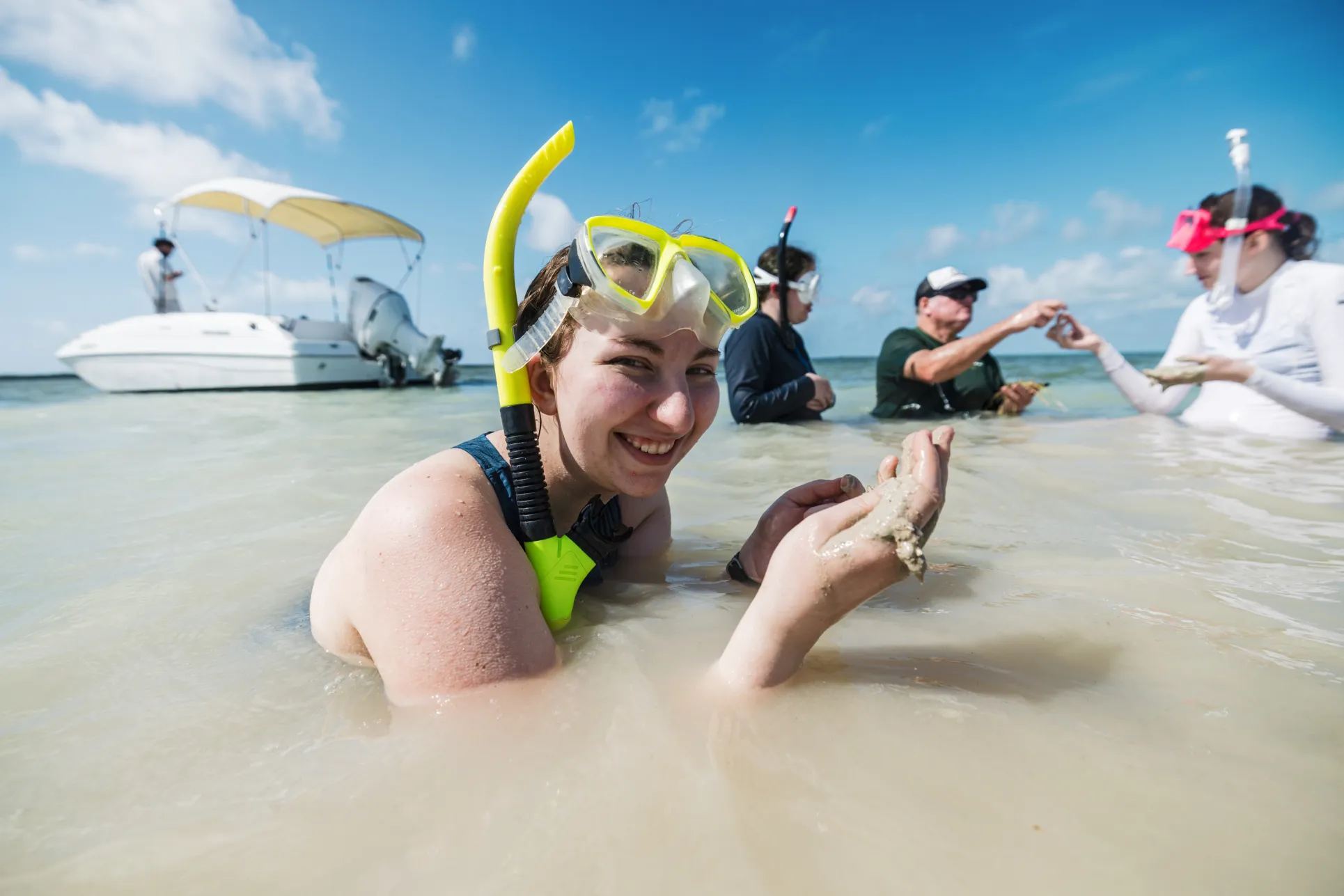 A young woman in a yellow snorkel and swim gear holds a sea life specimen while standing in muddy water and smiling at the camera
