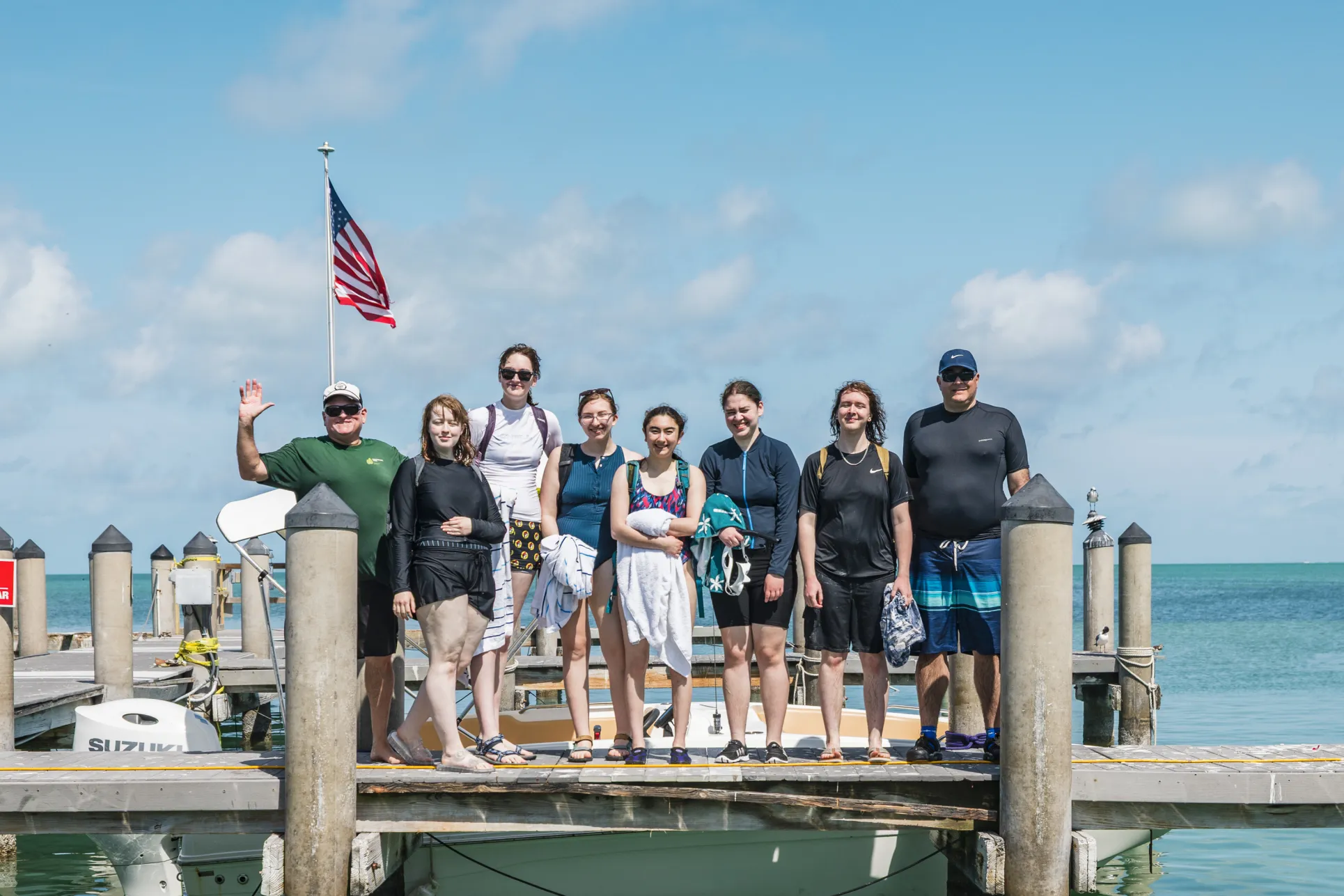 A team of students and professors in swimsuits and wetsuits stand on a dock with blue sky, clouds, and an American flag behind them and pale blue-green water below