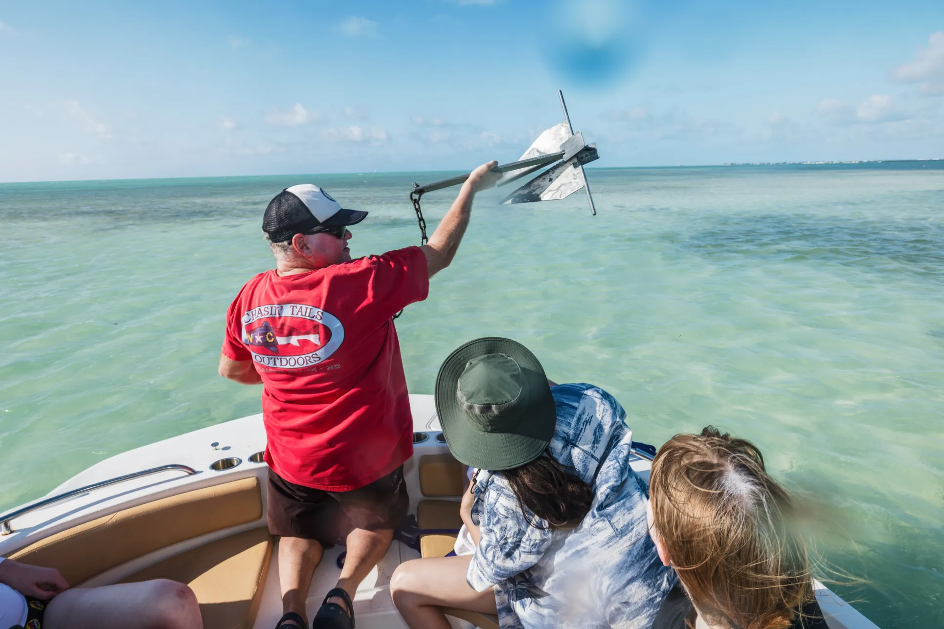 A man in a red shirt holds an anchor above the bow of a small research boat with two students looking on, as seen from behind