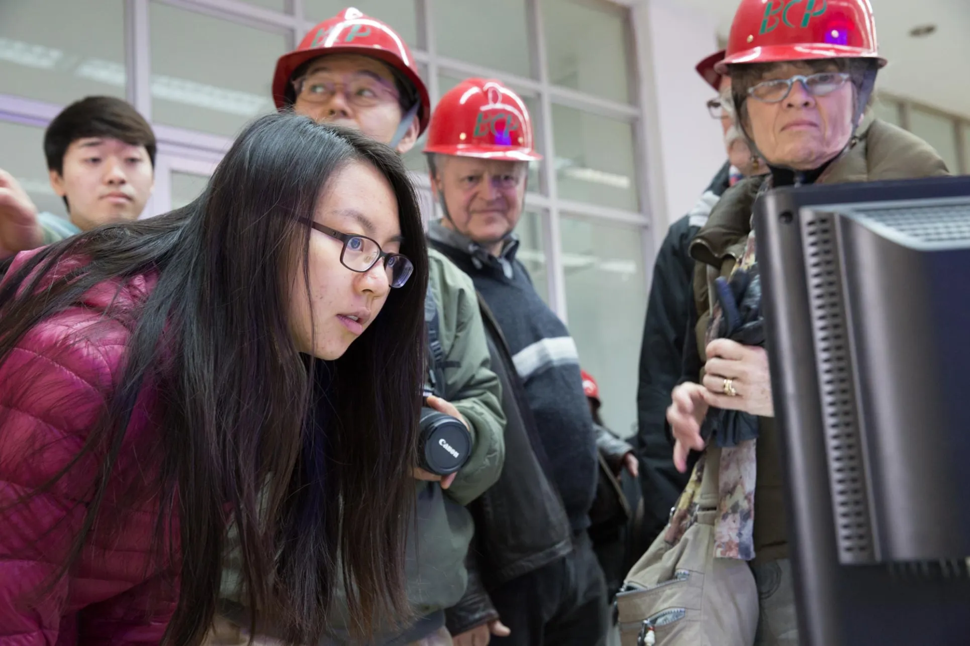 A young woman with long black hair and glasses looks at a computer screen while several people in red hard hats stand behind her 