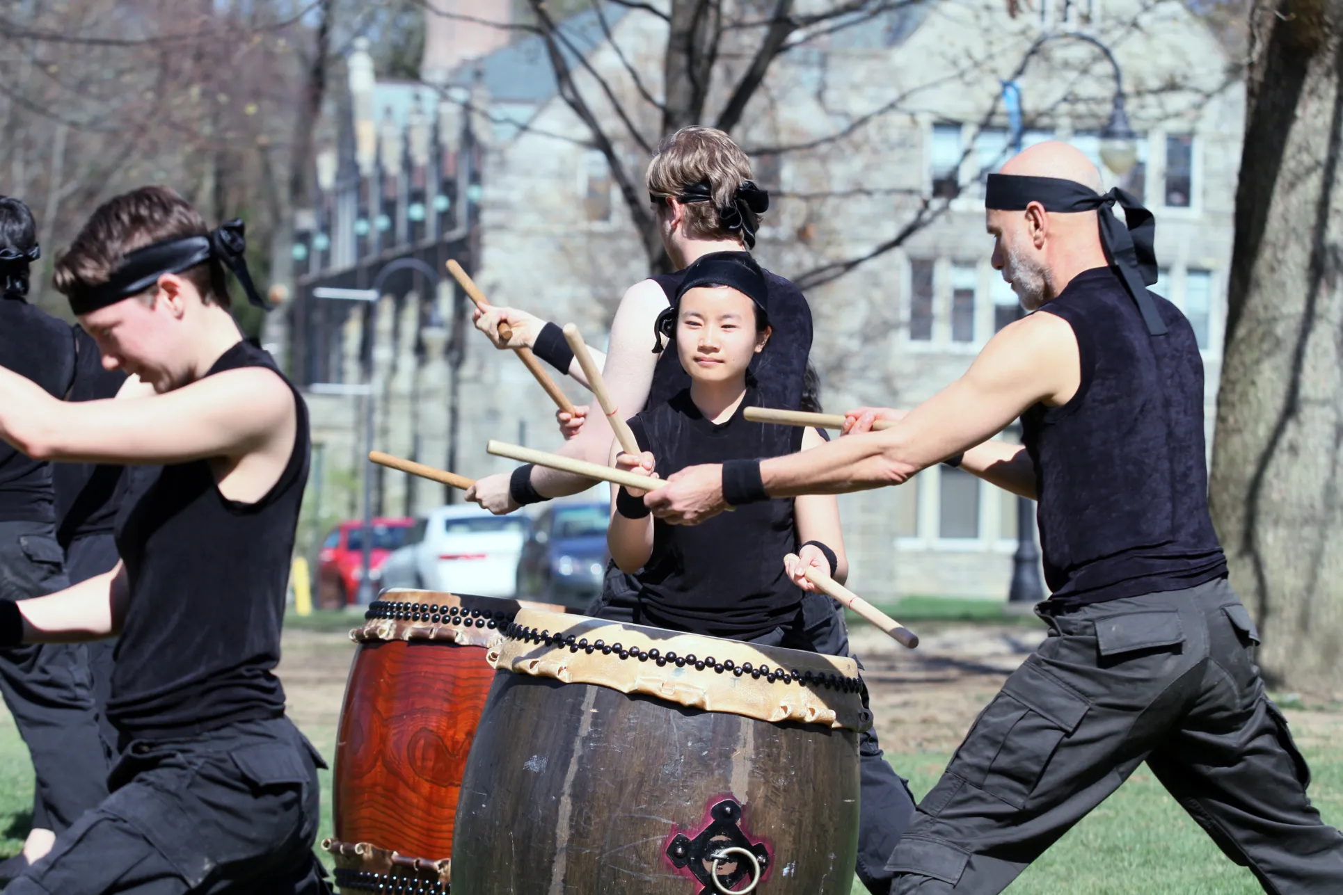 Four men in black pants and tank tops wearing black headbands perform with Japanese Taiko drums on a field with buildings behind them