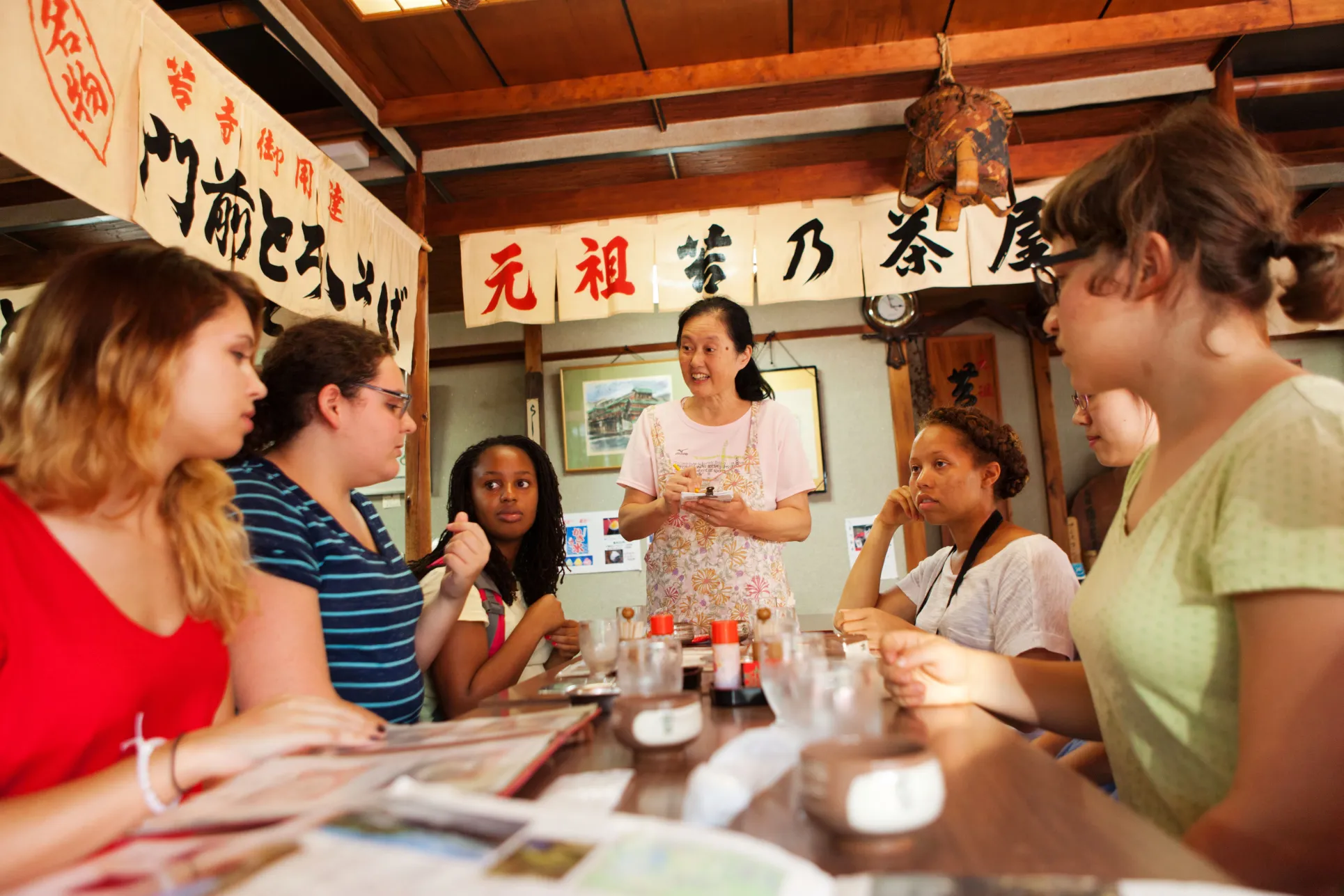 A group of six students sit around a table as a waitress takes their order. The walls have Japanese writing on small white sheets hanging near the ceiling.