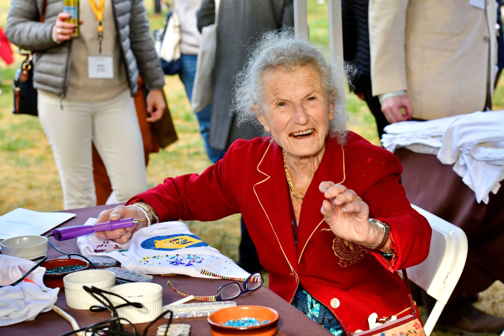 A woman in a red velvet school blazer smiles at the camera mid-craft. She is bedazzling an inauguration tshirt.