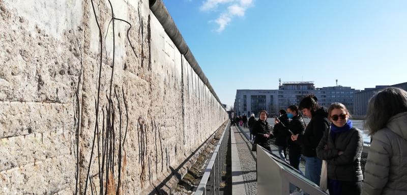 A stone wall with line drawings of figures on the left side of the frame; a row of 6 students and faculty on the right side of the frame observe and read a plaque about the art under a blue sky.