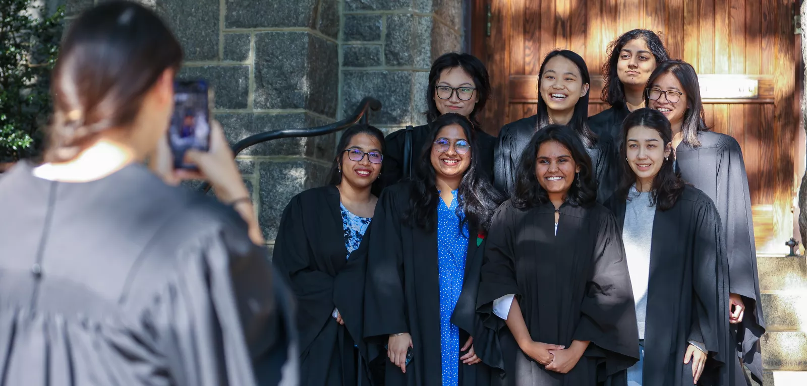Students wearing dark robes smiling on the Senior Steps as another student takes a photo of them on a phone