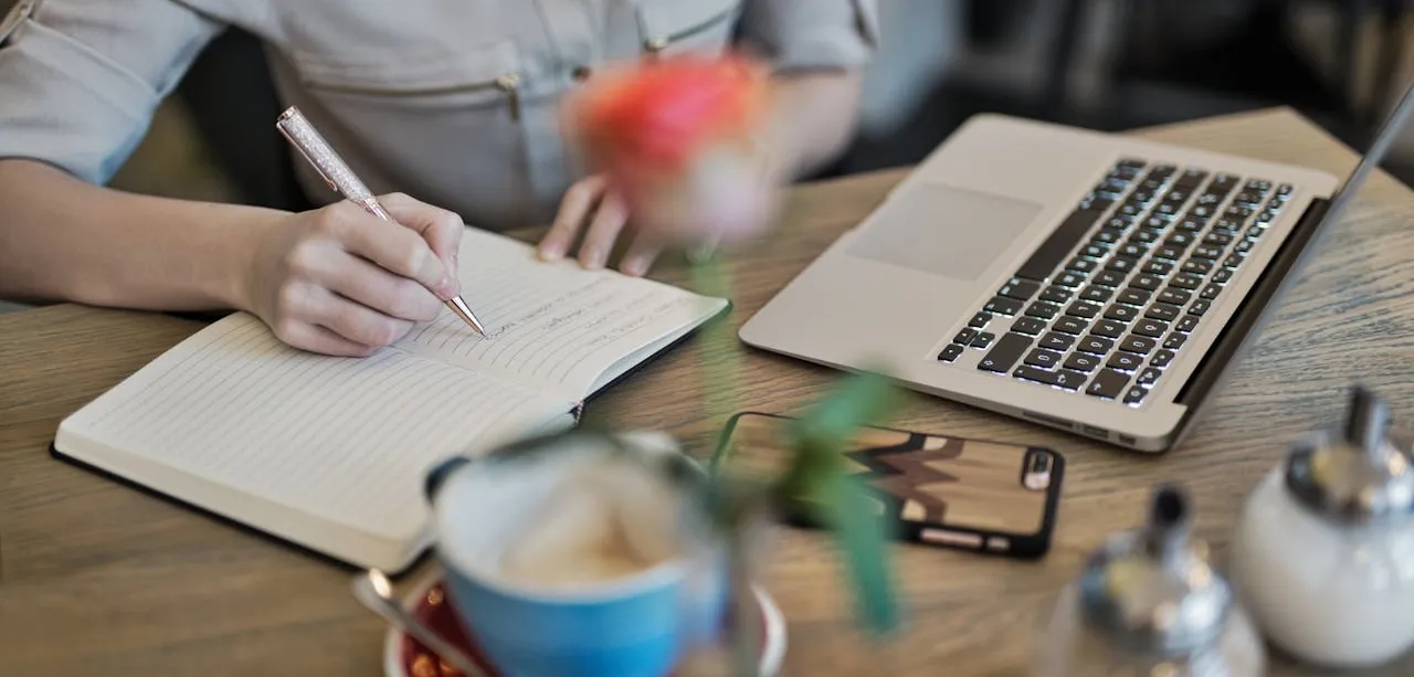 Person writing with a laptop, phone, coffee, and plant