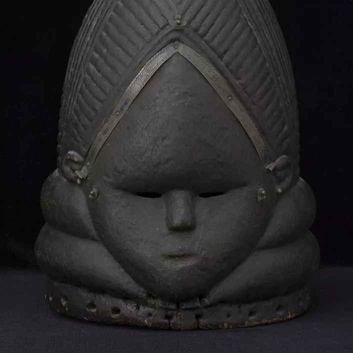 Blackened wooden mask of a diamond-shaped face, framed by a triangular hairline and chinline. The hairline is embellished with applied metal strips, above which is an elaborate coiffure of braids and bun.
