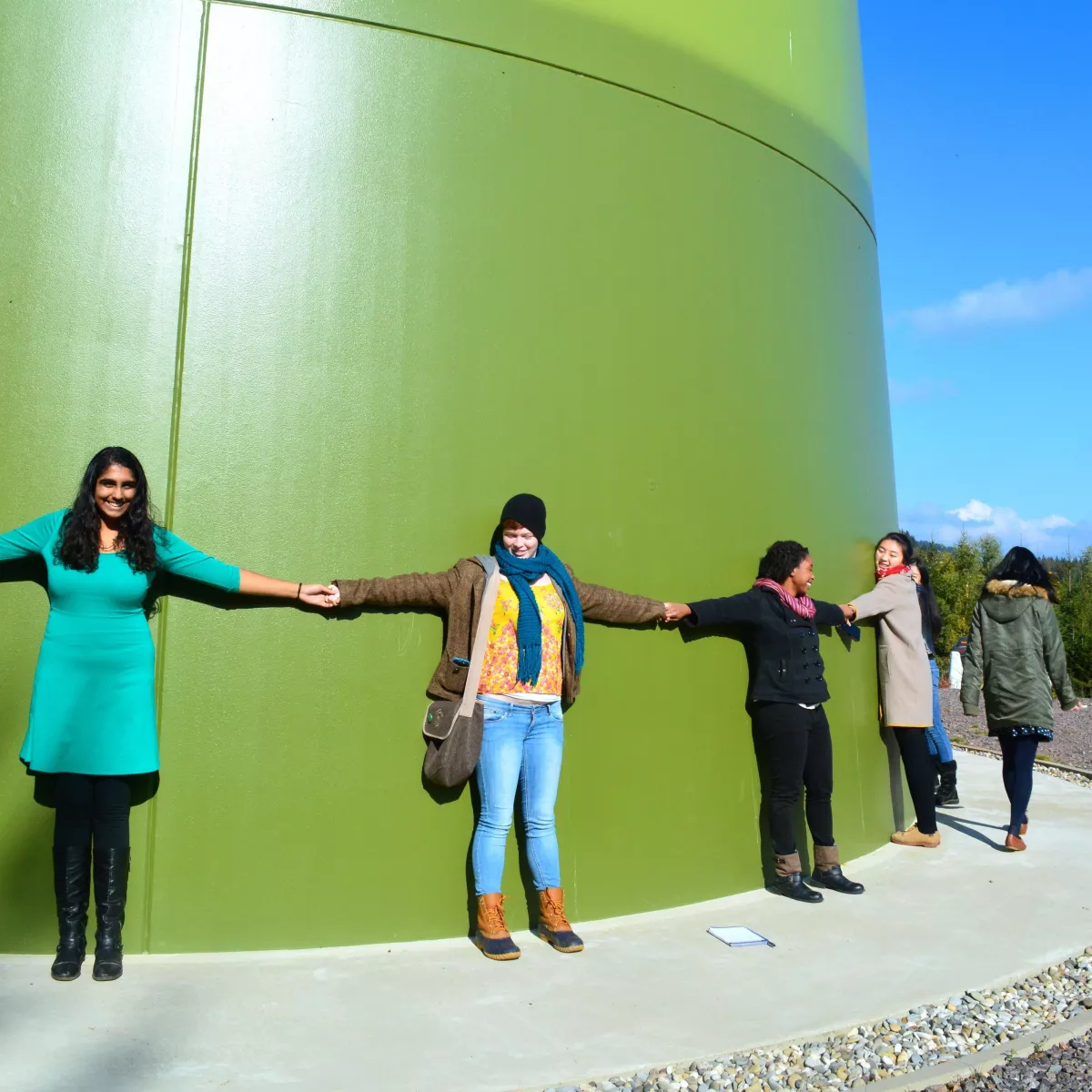 Students hold hands around the enormous green base of wind turbine.
