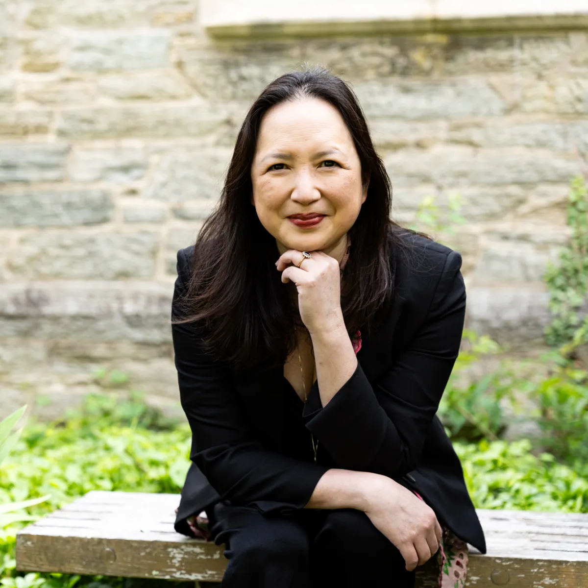 A woman with long straight dark hair and light skin wearing a black suit sits with her chin on her left fist. She is on a stone bench in front of a wall in a garden