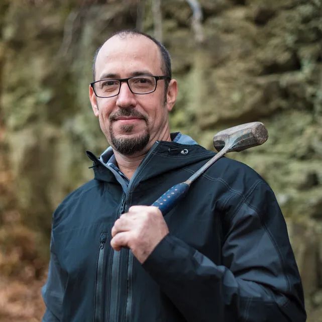 A balding man with light skin, a goatee and glasses wears a jacket standing outdoors with a mallet over his shoulder