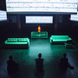 Three people sit on the floor across from three green couches on a dark stage. Four large projections of sheet music hang above the couches.