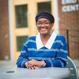 Saeina Charles smiling and seated at a table in front of the Enid Cook Center