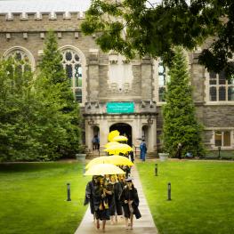 Class of 2023 walks out of Old Library wearing their cap and gown under yellow umbrellas